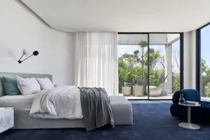 A minimalist bedroom featuring fabric bedding, glass and concrete surfaces, and a large window with a view of greenery outsid