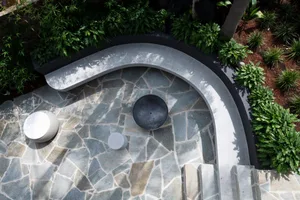 A modern patio featuring a curved granite bench and circular tables surrounded by green plants.