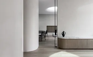 A minimalist dining room with wooden flooring, a wooden table, black chairs, and concrete walls.
