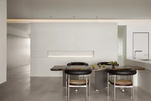 A modern minimalist kitchen with a wooden table and minimalist chairs in front of a concrete wall with recessed lighting. The