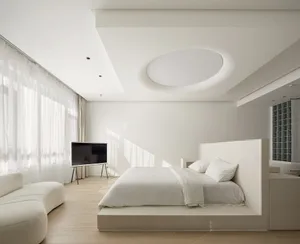 A minimalist bedroom featuring white furniture and a large window with sheer curtains.