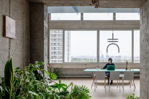A modern, concrete workspace area with a person working on a laptop, surrounded by large windows overlooking a cityscape.