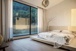 A minimalist bedroom featuring a wooden platform bed with white bedding, large windows, and a floor-to-ceiling window showcas