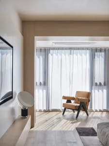 A minimalist living room with a wooden floor, a brown leather armchair, and sheer curtains allowing natural light to filter i