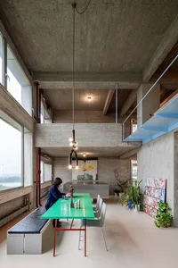A minimalist industrial-style loft with concrete walls, a green dining table, and blue stairs.