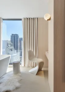 A modern minimalist bathroom with white decor, featuring a sleek white chair, a free-standing bathtub, a wooden stool, and a 