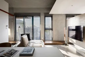 A modern living room with concrete walls and wooden accents, large windows, and a sleek sofa.