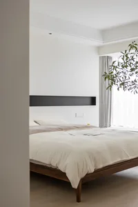 A minimalist bedroom with a wooden frame bed, white bedding, and a large potted plant by the window.