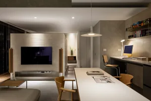 A minimalist living room with a concrete surface, featuring a wall-mounted TV, a long table with a computer setup, and a comf