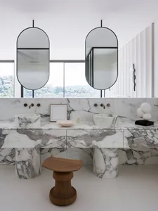 Modern bathroom with marble countertops and sinks, two oval mirrors, and a view of the city.