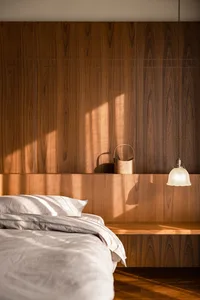 A bedroom with a wooden wall featuring a hanging lamp, a basket, and a neatly made bed with white bedding.