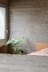 A minimalist bedroom with concrete walls, featuring a bed with a pink sheet and a green plant.