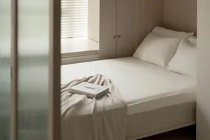 A minimalist bedroom with a white bed featuring a book on the bedspread, surrounded by neutral tones and soft lighting.
