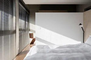 A minimalist bedroom with a white bed and a modern chair next to a large window with blinds.