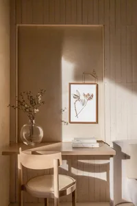 A minimalist workspace featuring a wooden desk, chair, vase with dried flowers, and a framed botanical print on the wall, wit