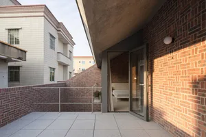 A modern residential balcony with a brick wall on the left and concrete flooring. The interior reveals a minimalist bedroom s