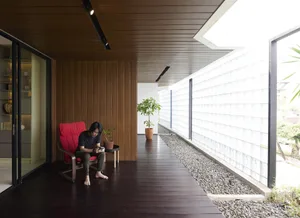 A person sits on a red cushioned chair in a modern porch space with a striped wooden floor, green plant in a terracotta pot, 