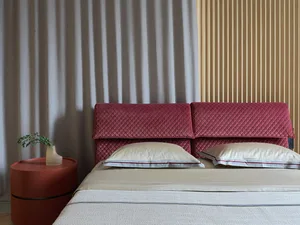 A minimalist bedroom featuring a red velvet headboard and white bedsheets on a bed with a round red bedside table holding a v