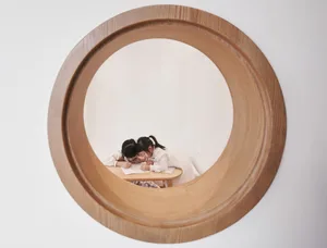 Two children sitting at a wooden table in a classroom, viewed through a round wooden window.