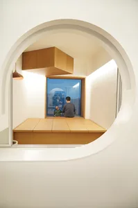 現代室內空間，設有圓形窗戶、木製長椅，以及一位大人與小孩一同坐在長椅上。