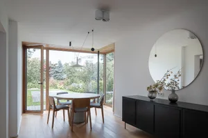 A modern minimalist dining area with a round table, chairs, and a large window overlooking a garden.