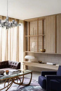 A modern living room featuring a natural wood shelf, a glass coffee table, and two armchairs, with a chandelier hanging from 