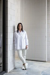 A woman wearing a white button-down shirt and white pants standing in front of a white wall and vertical blinds.