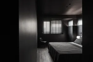 Minimalist bedroom with wood flooring and furniture, featuring a single window with blinds and a lit lightbulb.