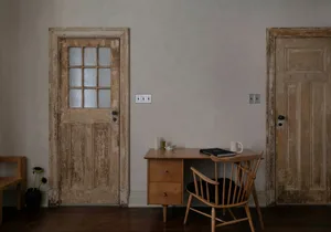 A minimalist interior featuring a wooden desk and chair against a plain wall, with two doors in the background.