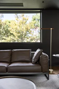 A modern minimalist lounge with a brown leather sofa, wooden floor lamp, and large window overlooking trees.