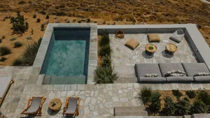 A minimalist outdoor living space featuring stone pavers and concrete, surrounded by natural vegetation and a small infinity 