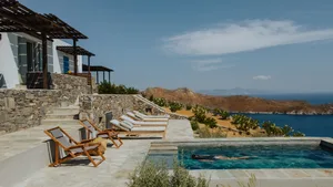 Stone and wooden pool deck with concrete stairs, glass railing, and timber lounge chairs overlooking the ocean.
