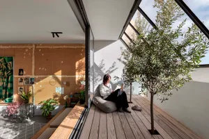 A woman sits on a cushioned chair reading a paper in a modern living space with wooden flooring and plants.