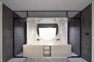 A modern bathroom interior featuring a marble countertop and shower area with a large round mirror and minimalist fixtures.