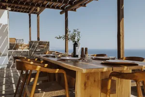 A wooden outdoor dining table set with plates, glasses, and a bottle of wine, overlooking the sea.