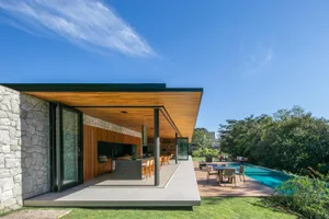 A modern residential patio with a wooden roof and glass walls, surrounded by greenery and featuring outdoor furniture.
