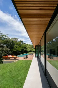 Modern house exterior with wood accents, concrete patio, and glass windows overlooking a pool and lush greenery.
