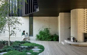 A modern house exterior with a combination of concrete walls and stone elements, surrounded by greenery and a wooden deck.