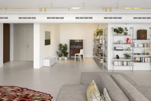A modern living room with a concrete floor, featuring a gray sectional sofa, bookshelves, and potted plants.