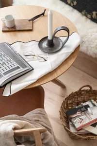 A minimalist living room reading corner featuring candle holder, glasses, books, and wooden furniture on a round wooden table