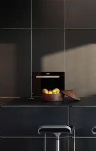 A modern kitchen interior featuring sleek, black cabinets and a countertop, with a wooden bowl containing a variety of fruits