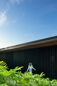 A woman walks beside a modern house with black vertical bars and green foliage in the foreground.