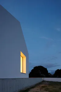 A modern white house with geometric concrete cuts, seen at dusk with illuminated windows.