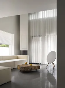 A modern, minimalist living room with white furniture, a wooden coffee table, and sheer curtains.