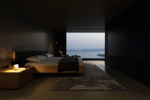 A minimalist bedroom featuring a wooden bed, a rug, and a view of a calm body of water.