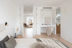 A minimalist bedroom with wooden floors and a bed dressed in neutral tones, featuring a study area and an open layout.