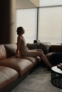 A woman in a sleeveless beige dress sitting on a modern leather sofa in a contemporary relaxation space with blinds.