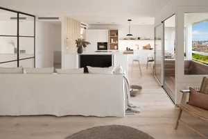 A modern living room with a white interior, featuring a large sofa, a kitchen island, and a balcony with a view of the city.