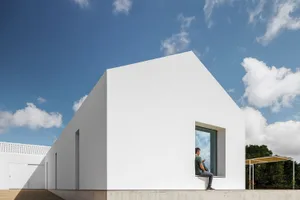 Man sitting on a modern minimalist white stucco residential building exterior under a blue sky with clouds.