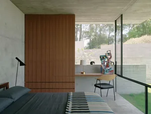 Modern minimalist bedroom featuring concrete walls and floor, with a wooden wardrobe and a glass desk with a colorful sculptu
